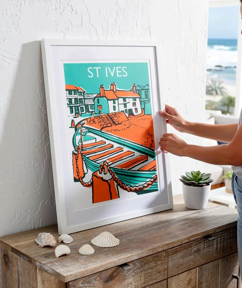 Framed St Ives boat-themed art print being positioned on a wooden surface, with seashells and a small plant nearby.